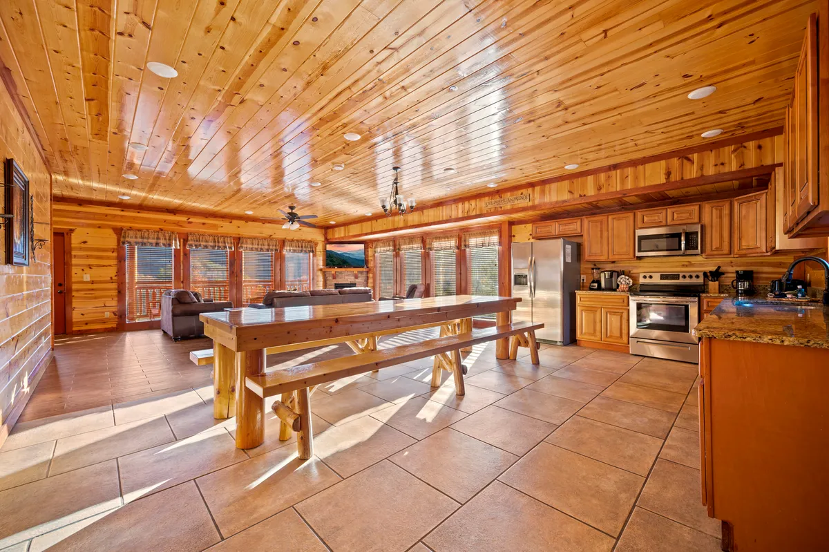 A spacious dining area with a large wooden table surrounded by ample seating, adjacent to a kitchen and living area. Floor-to-ceiling windows provide scenic views, while rustic finishes evoke a welcoming lodge atmosphere—ideal for shared meals and celebrations.