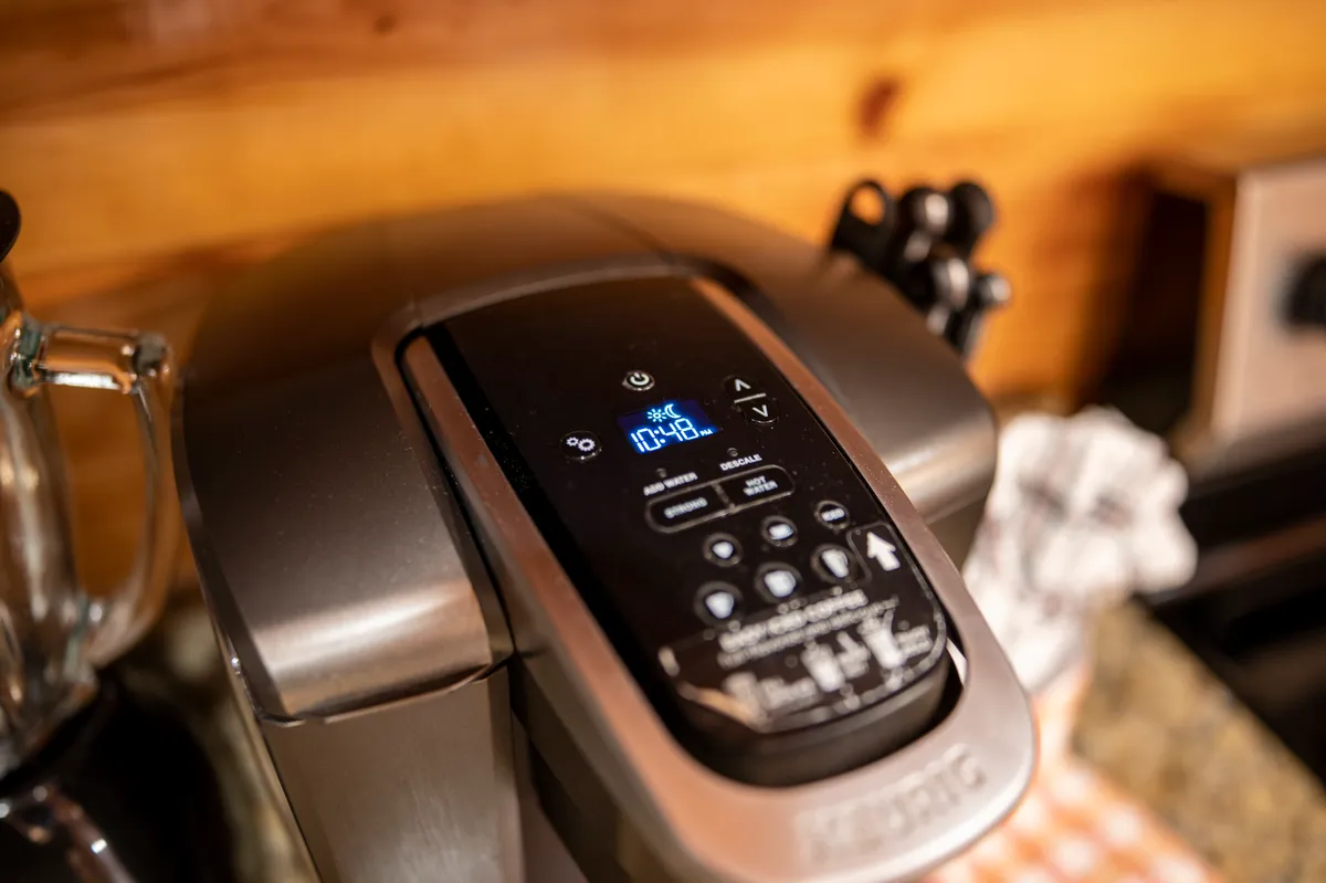 A cozy kitchen corner with a coffee maker set against rustic wooden walls and utensils neatly arranged, bathed in warm morning light. The setup suggests a space designed for both function and daily rituals.