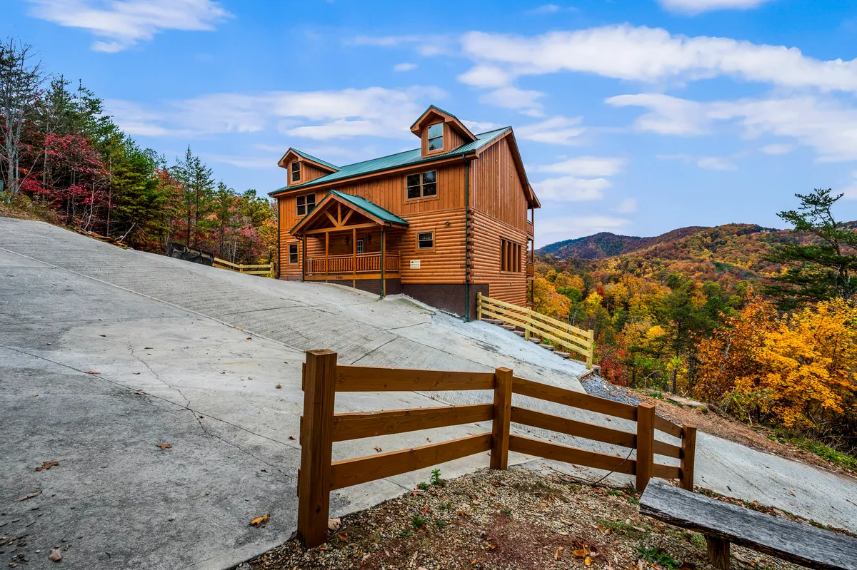 A cozy log cabin surrounded by mountains and colorful foliage, with a driveway leading to the entrance. The environment suggests easy access to hiking, nature walks, and outdoor gatherings, making it an inviting base for both guests and their pets.