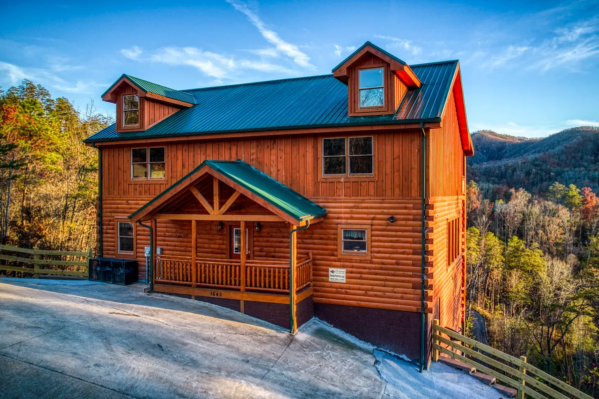 The image showcases the welcoming wooden exterior of a spacious cabin with a striking green metal roof, set against the scenic backdrop of the Smoky Mountains. The inviting entrance porch basks in the soft glow of sunset, hinting at cozy evenings and memorable family gatherings.
