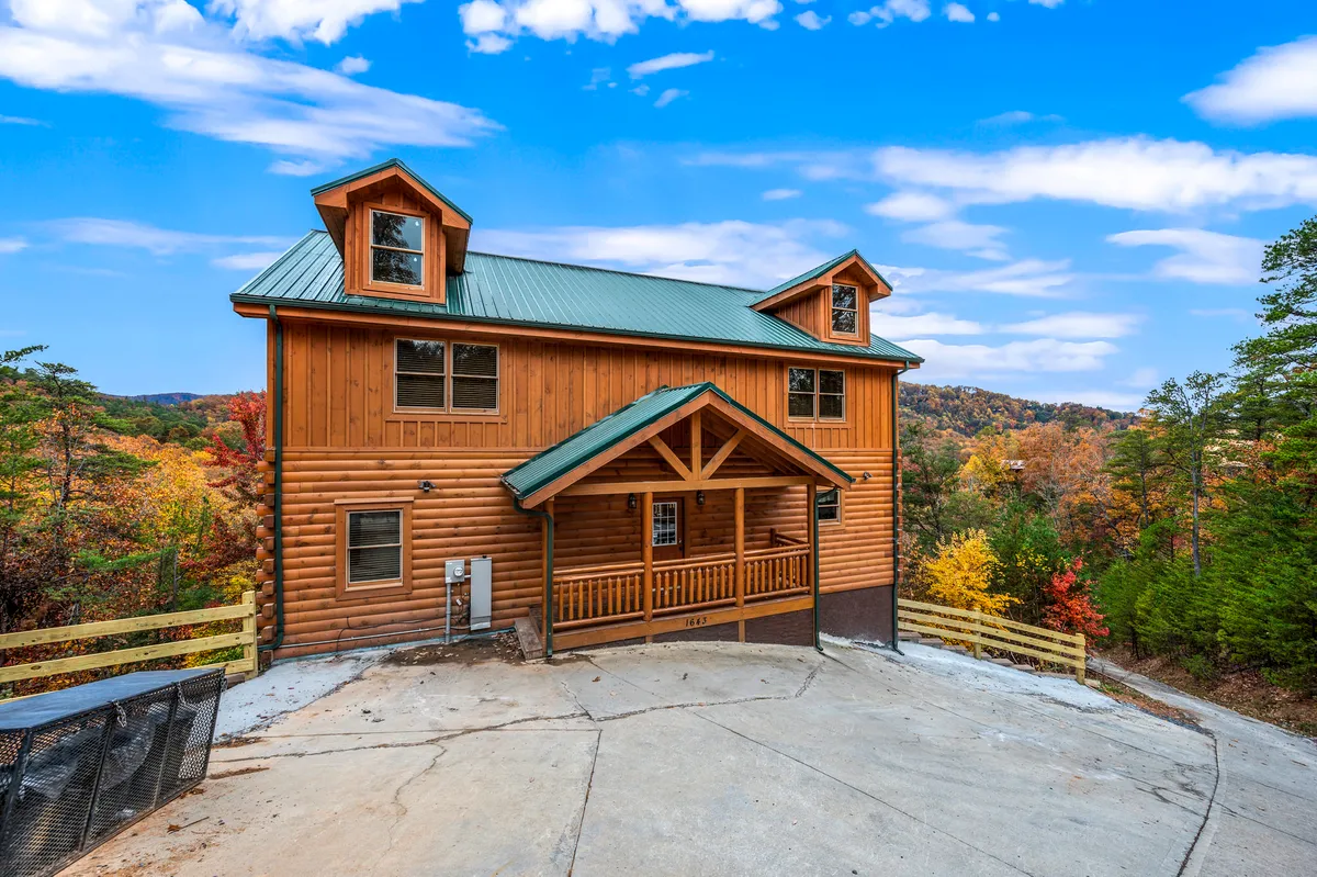 A welcoming log cabin with a green metal roof, set against a backdrop of colorful autumn foliage. The spacious porch with a wooden railing hints at a perfect spot for family gatherings, while the surrounding trees evoke a sense of adventure and tranquility. The cabin is nestled in nature, inviting exploration of nearby trails.