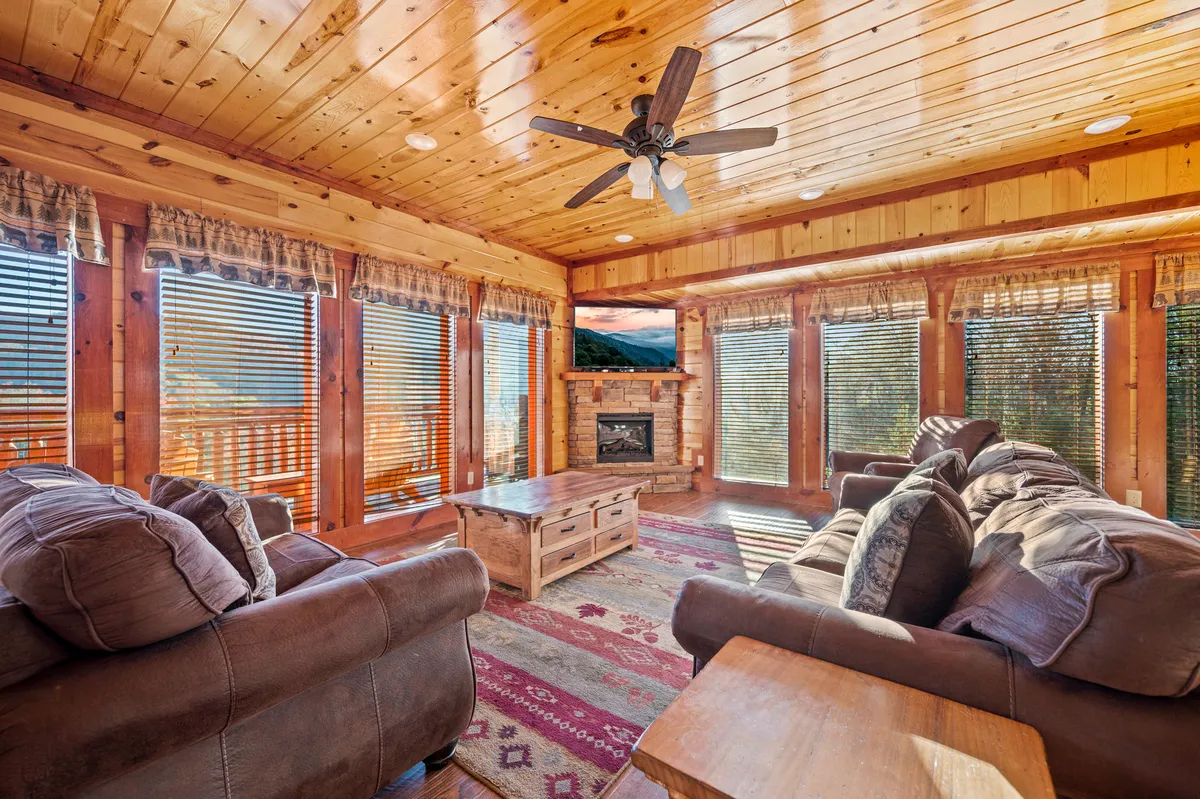 A spacious living room with large windows offering mountain views, a stone fireplace as a central feature, comfortable plush seating arranged for group gatherings, and rustic wooden accents throughout. Natural light floods the room, highlighting the inviting open-plan design.