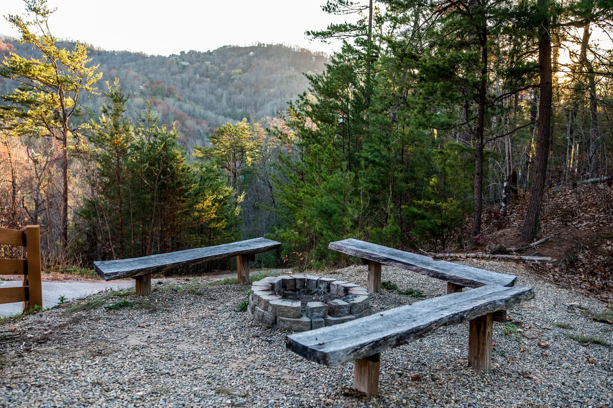 A fire pit surrounded by wooden benches and framed by tall trees, creating an inviting outdoor space for family and friends to gather. The setting is ideal for storytelling, roasting marshmallows, and enjoying the natural beauty of the mountains at sunset or under the stars.