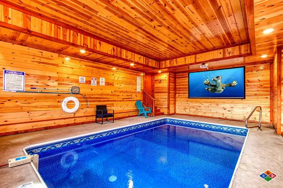 A vibrant indoor swimming pool features a large screen TV mounted on the wall, wood paneling for a cozy cabin vibe, and a comfortable lounge chair nearby. The pool area exudes a leisurely atmosphere with plenty of space for both swimming and relaxing, suggesting fun for groups of all ages.