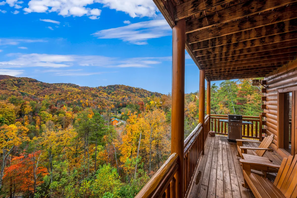 A spacious wooden deck with comfortable seating overlooks the Smoky Mountains, framed by rich fall foliage. The deck is bathed in natural light, ideal for relaxing after a day outdoors. The tranquil mountain view invites both humans and dogs to unwind in nature’s embrace.