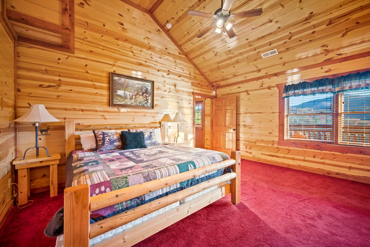 A warmly lit bedroom featuring a cozy wooden bed frame with a colorful quilt, inviting wooden walls, and large windows that offer scenic mountain views. The sunlight streaming in creates a peaceful and comfortable ambiance, perfect for a restful retreat.