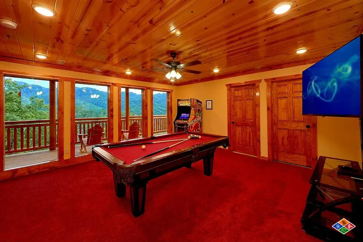 A lively game and arcade room equipped with a pool table, an arcade game machine, a television, and expansive windows framing mountain views. The space is bright and playful, designed for friendly competition and group fun while still celebrating the cabin's scenic surroundings.