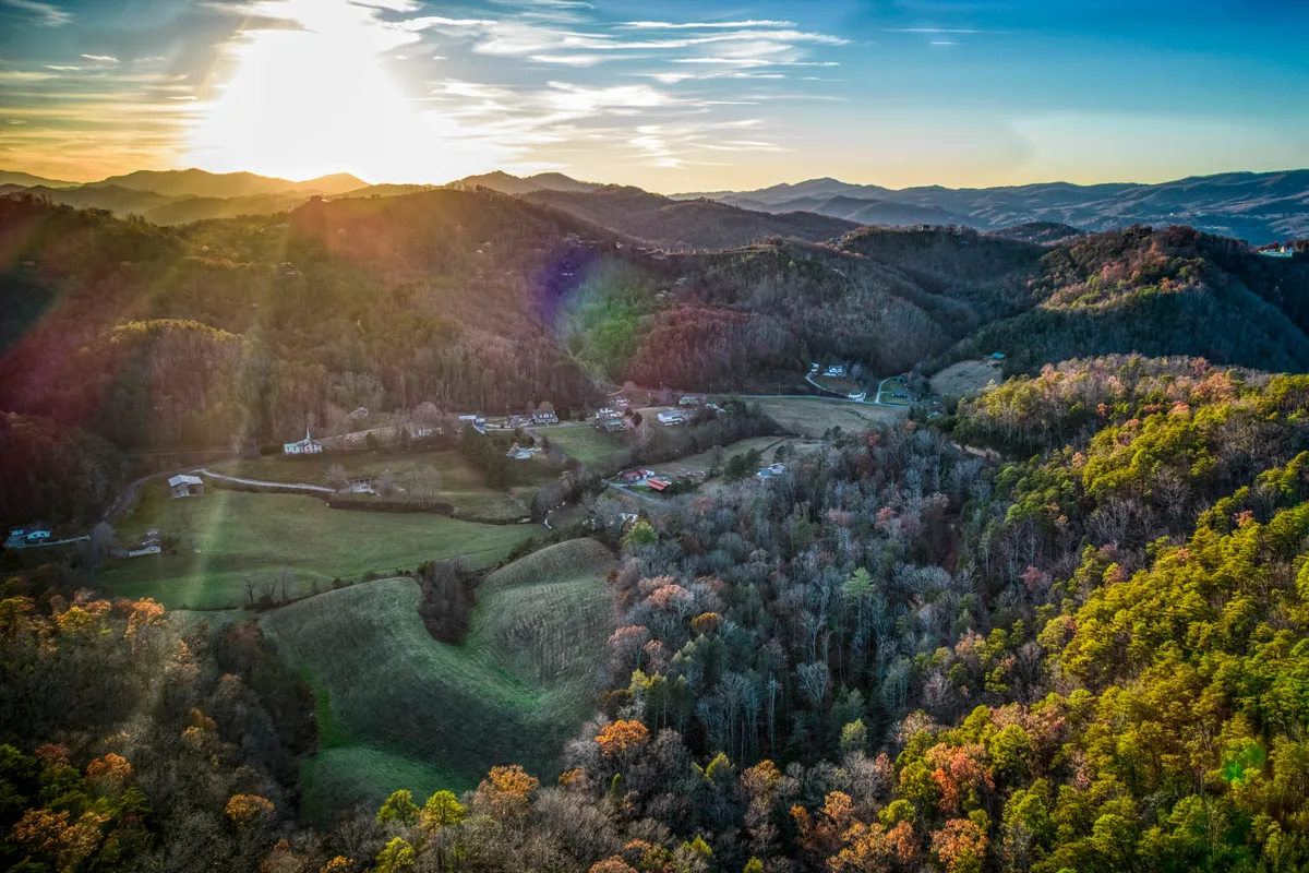 A panoramic view of rolling hills and distant mountains, bathed in the warm hues of sunset. Autumn foliage creates a rich tapestry of color, with rural homes dotting the landscape. The tranquil scene evokes a sense of peace and awe-inspiring natural beauty.